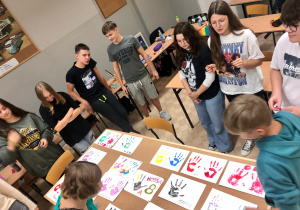 Zabawy integracyjne w klasie 8A. Uczniowie stają w kręgu przy stole i ogladają swoje prace, czyli kolorowe odciski swoich dłoni na białym papierze, przyczepione do korkowej tablicy.